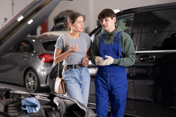Young guy mechanic advises young woman client on repairing under hood of car in car service station