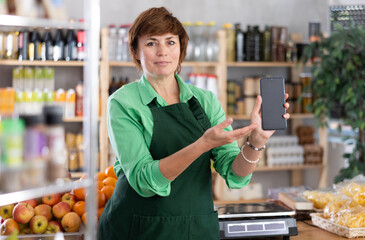 Woman seller stand shop, shows smartphone screen, points to device and offers convenient way for...