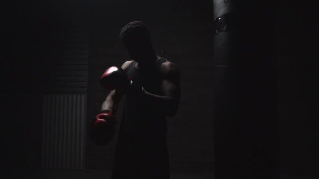 african american athletic man putting on red boxing gloves in a dark room, strong boxer preparing for training and fight