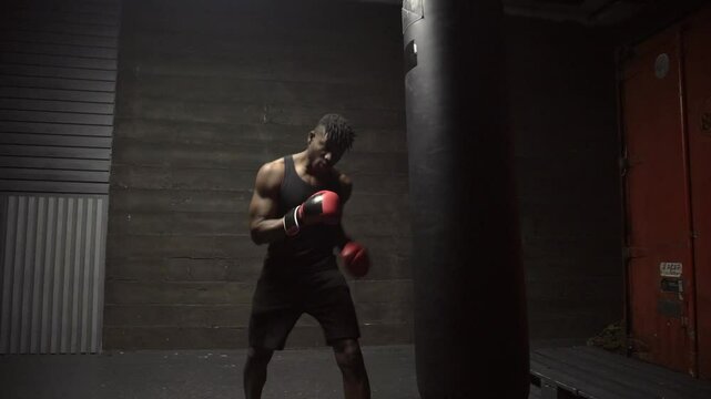african american young boxer training in dark gym and hitting punching bag with gloves, strong man doing sports and training endurance