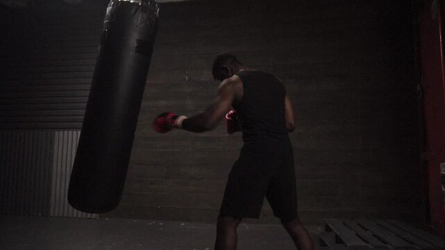 african american young boxer training in dark gym and hitting punching bag with gloves, strong man doing sports and training endurance