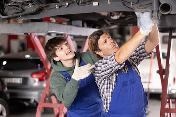 Young guy and adult male mechanic in uniform repairing underbody of car in car service station