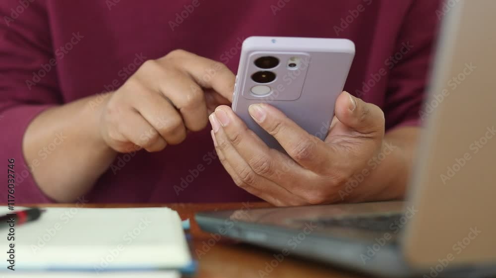 Poster Closeup of man hand using mobile phone while working on laptop computer at home. Male fingers typing and tapping on smartphone. Texting and scrolling.