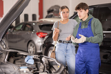 Young guy mechanic advises young woman client on repairing under hood of car in car service station