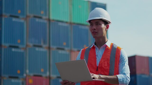 A Southeast Asian males wearing a hard hat and an safety vest, speaking into a walkie-talkie and checking data in laptop computer at container yard. They 'll ship containers in Logistic area.