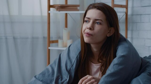 A millennial woman sits wrapped in a blanket, eating a snack, while looking contemplative and disconnected. The atmosphere reflects feelings of depression and loneliness during winter.
