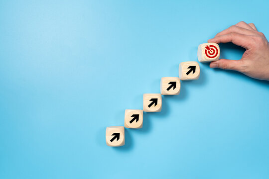 Wooden blocks forming a staircase with upward arrows, leading to a target symbol, symbolizing progress and goal achievement against a light blue background.