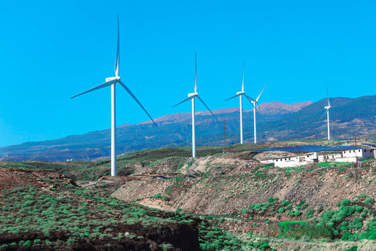 Series of wind turbines on a hilly landscape with a clear blue sky in the background. There are also a few buildings visible near turbines, indicating a wind farm setup for generating renewable energy