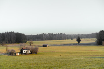 Wide view of a serene rural countryside featuring a small white cottage, open grassy fields, and a distant forest.