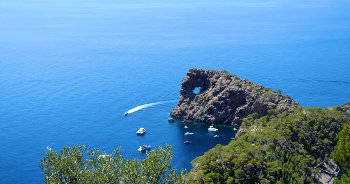 Vivid 4K DCI footage showcasing the bright blue waters surrounding Sa Foradada, with lush green trees framing the iconic rock formation and anchored boats below.--