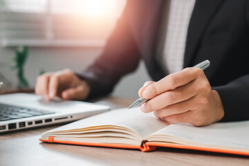 Close-up of a business professional writing in a notebook at a desk with a laptop, illuminated by soft sunlight.