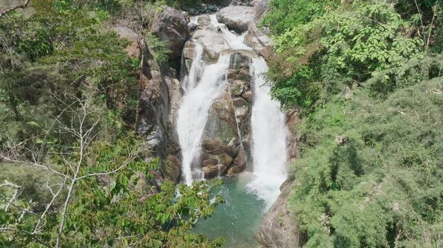 Majestic tropical waterfall cascades down rugged granite cliffs in Costa Rica (San Gerardo de Rivas). Drone shot reveals dual streams merging into turquoise pool, surrounded by lush rainforest.