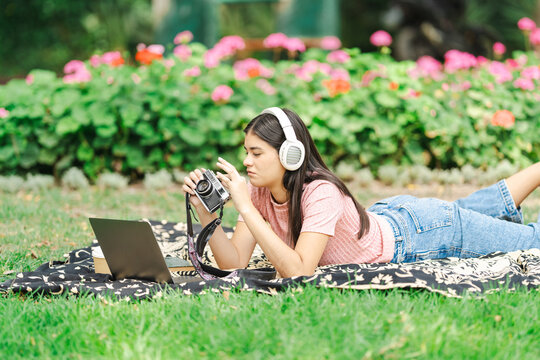 Young photographer reviewing photos on vintage camera while lying in park with laptop and book