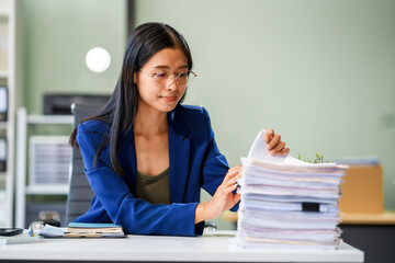 A businesswoman sits at her desk in a modern office, surrounded by numerous documents. She works...