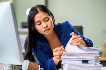 A businesswoman sits at her desk in a modern office, surrounded by numerous documents. She works...