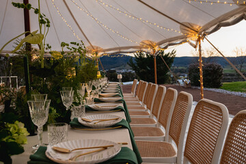 A beautifully arranged outdoor dining setup under a white marquee tent. The table is adorned with lush greenery, delicate floral arrangements, and gold-accented cutlery, paired with crystal-clear glas