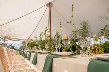 A beautifully arranged outdoor dining setup under a white marquee tent. The table is adorned with lush greenery, delicate floral arrangements, and gold-accented cutlery, paired with crystal-clear glas