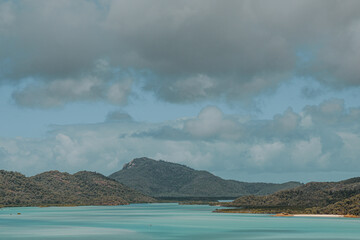 A stunning panoramic view of a tropical beach with crystal-clear waters and white sand, surrounded by lush greenery and rolling hills. The sky is partially cloudy, adding depth to the scenery, while s