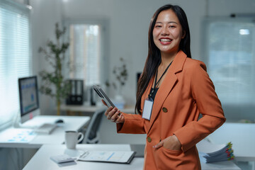 Young manager in an orange suit, wearing a lanyard and badge, holding a smartphone while standing confidently in a bright office with a hand casually placed in her pocket