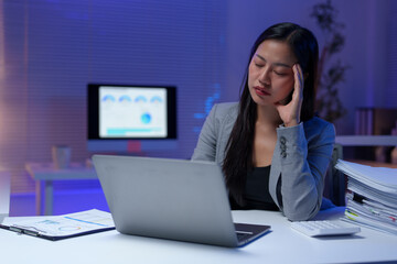 Stressed and tired businesswoman having a headache while working late at night in office using laptop, with financial reports and graphs showing on computer screen