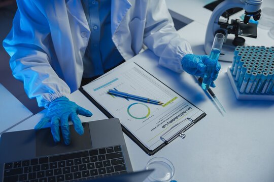 Scientist wearing lab coat and gloves analyzing data on laptop and holding test tube with blue liquid, surrounded by lab equipment and reports in modern laboratory