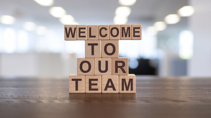 Four wooden blocks stacked vertically display "Welcome to our team" in dark sans-serif font. Set on a dark surface, the blurred bright background evokes a professional, welcoming office ambiance.




