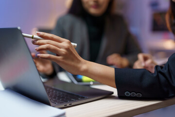 Businesswomen collaborating over a laptop and digital pen during a nighttime meeting in the office, focusing on hands while the background remains softly blurred