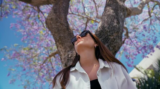 Close-up of a young adult woman sits beneath a jacaranda tree adorned with vibrant purple flowers, basking in the sunlight