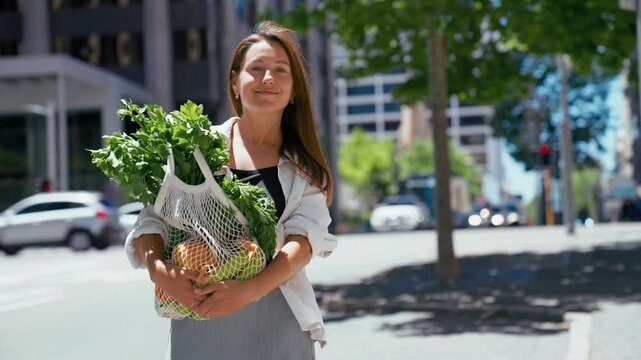 Portrait of a young woman smiles while carrying a reusable bag filled with fresh fruits and vegetables on a sunny urban street.