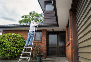 Man standing on the ladder and cleaning house with a long brush. Home maintenance work. Auckland. © Janice