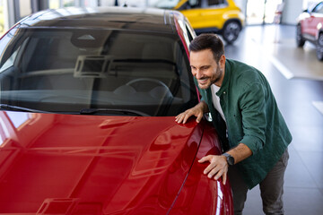 Handsome man buying new car, standing at car salon and checking automobile. Smiling guy looking at...