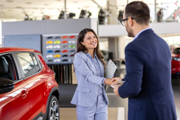 Smiling salesman or manager giving car keys and shakes hand to modern happy woman new car owner in car dealership