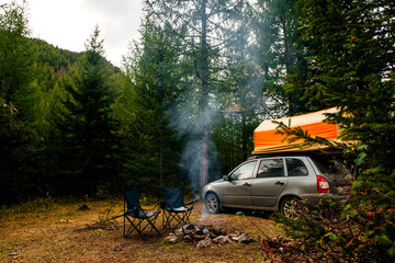a car with a tent on the roof in the forest, two chairs by the campfire