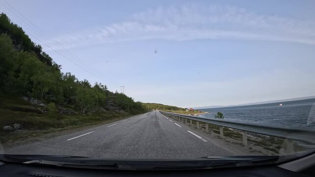 Driving along Norway's northern taiga on E69 highway. Low trees and rocks line the left side, while the sea is on the right. A few scattered houses add charm to the scenic landscape.