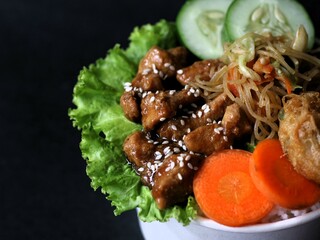 Teriyaki glazed chicken rice bowl with vegetables, noodle, sliced cucumber and wortel and mashed potatoes on black background. 