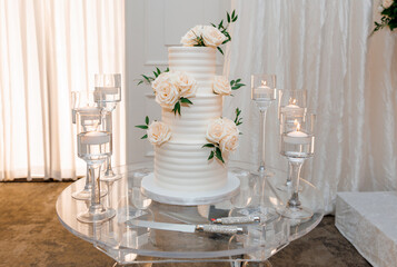 Elegant white three-tiered wedding cake with fresh white roses. A creamy vanilla wedding cake stands on a glass table and candles in glasses with water around. Festive cake for guests at the wedding