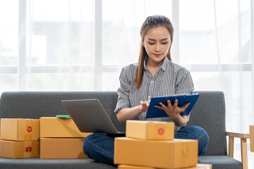 Online Business Owner Managing Orders: A young Asian woman sits comfortably on a sofa amidst a pile of shipping boxes, meticulously reviewing orders on a clipboard and laptop.