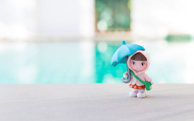 Miniature girl holding blue umbrella on swimming pool edge with space on blurred background, rainy season concept, school girl in raincoat