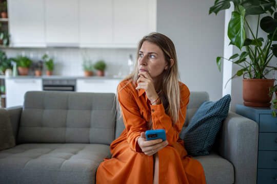 Thoughtful serious woman waits for message or call on phone, sitting alone on couch at home. Female quiet anticipation, solitude, waiting for connection, stress and overthinking, bad news, sorrow