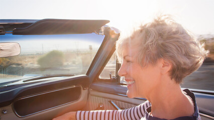 Senior Woman Passenger On Road Trip Driving In Classic Convertible Car Along Country Road