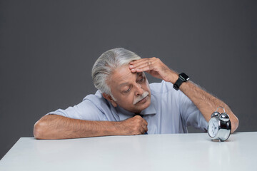Senior Finance Manager Hand On His Head And Is Looking At An Alarm Clock.�