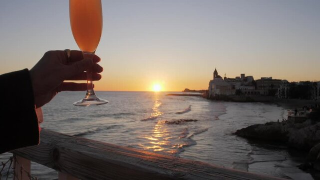 Glass of orange Bellini cocktail in woman hand, with sea and waves in background during golden hour. Romantic sunset date at cafe in historic European town Sitges.