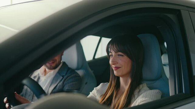 Happy Caucasian woman sitting in car and gently touching steering wheel. Inspective every detail of new vehicle. Shopping in dealership with boyfriend. Looking at camera while smiling.