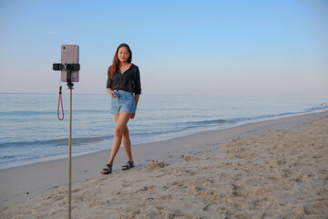 Beautiful asian woman standing on the beach using a selfie stick and smartphone to take portrait photos by the sea and blue sky background with copy space