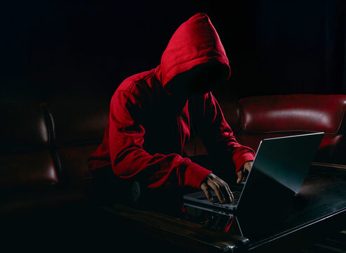 Hackers wear red hoody to cover her faces and sitting in the red sofa dark room. Hacking steal important information using laptop computer to release malware viruses. Ransom and harass organizations.