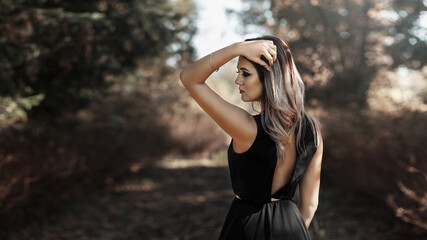 woman with long blond hair standing on a forest path. She is wearing a black dress with an open back. The woman stands sideways to the camera, raising one hand and placing it on her head.