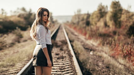 woman standing on the railway tracks. She is wearing a white shirt and a short black skirt. She has long blonde hair that falls loosely to her shoulders.