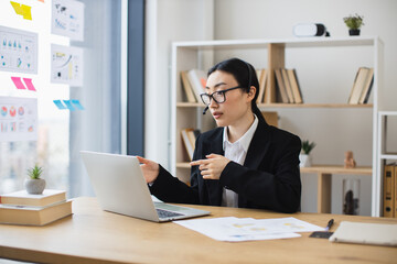 Asian businesswoman in black suit using laptop with headset during video call. Young professional...
