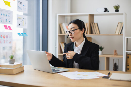 Asian businesswoman in black suit using laptop with headset during video call. Young professional engaging in online meeting in bright modern office setting with bookshelf and plant.