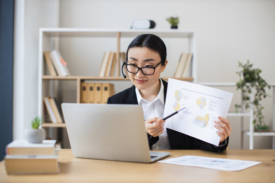 Confident Asian businesswoman wearing headset presenting data during video call using laptop. Middle-aged woman working remotely in office setting showcasing professional skills and communication.